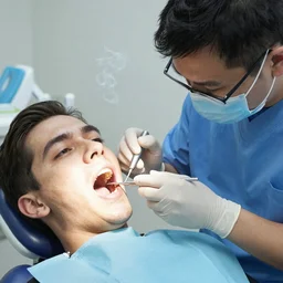 The dentist is drilling a tooth in a patient's mouth while smoking a cigarette held by his assistant