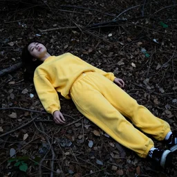 young female wearing a yellow velor sweat suit lying dead on the ground in a forest covered by dark colored leaves and twigs