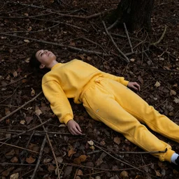 ”female laying down in forest covered by dark colored leaves and twigs wearing yellow velor sweat suit