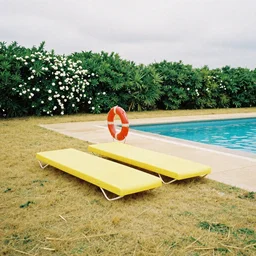 two yellow beach beds and a red life saver next to a pool, yellowish grass, green bushes in the background with white flowers, grainy photograph, diagonal composition, cloudy day, long shot