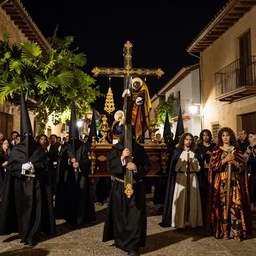 A Holy Week procession of souls in a lush Spanish village at night. A figure at the front holds a cross. El Greco, Remedios Varo y Salvador Dali.