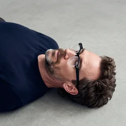 bryan cranston with a black clear reading glasses, grown hair on top of his head and a beard, laying on his back at smooth concrete floor, digital art